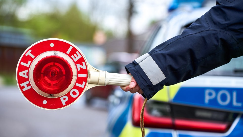 Der Arm einer Polizistin vor einem Polizeiauto mit Polizeikelle an einer Straße