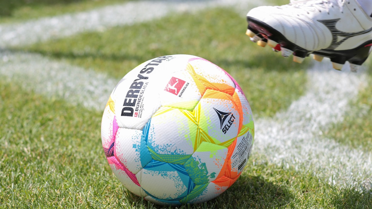 Derbystar Fußball mit Logo der 1. Bundesliga der Männer auf dem Rasen. Ein Fußballerbein im Anschnitt.